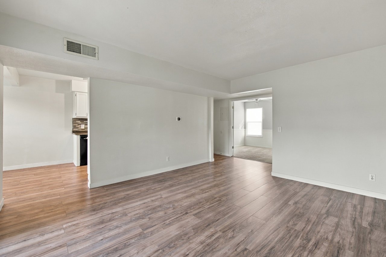 Vacant living room with wood-look flooring and open doorways to the kitchen and adjoining room.