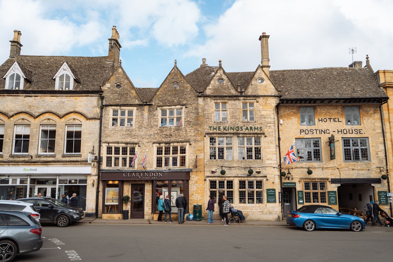 Historic stone village street with the Kings Arms pub, shops, pedestrians, and parked cars.
