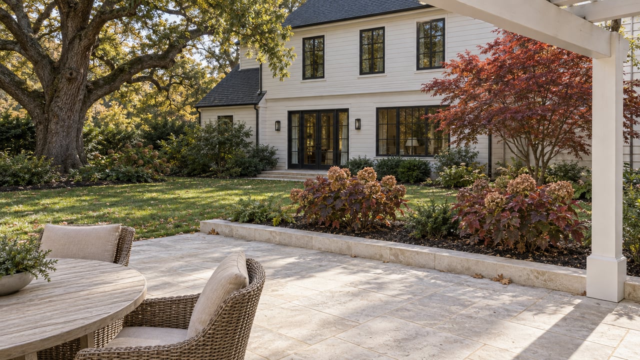 Limestone garden terrace beside an ivory-sided home, with black divided-light windows, woven chairs and autumn foliage.