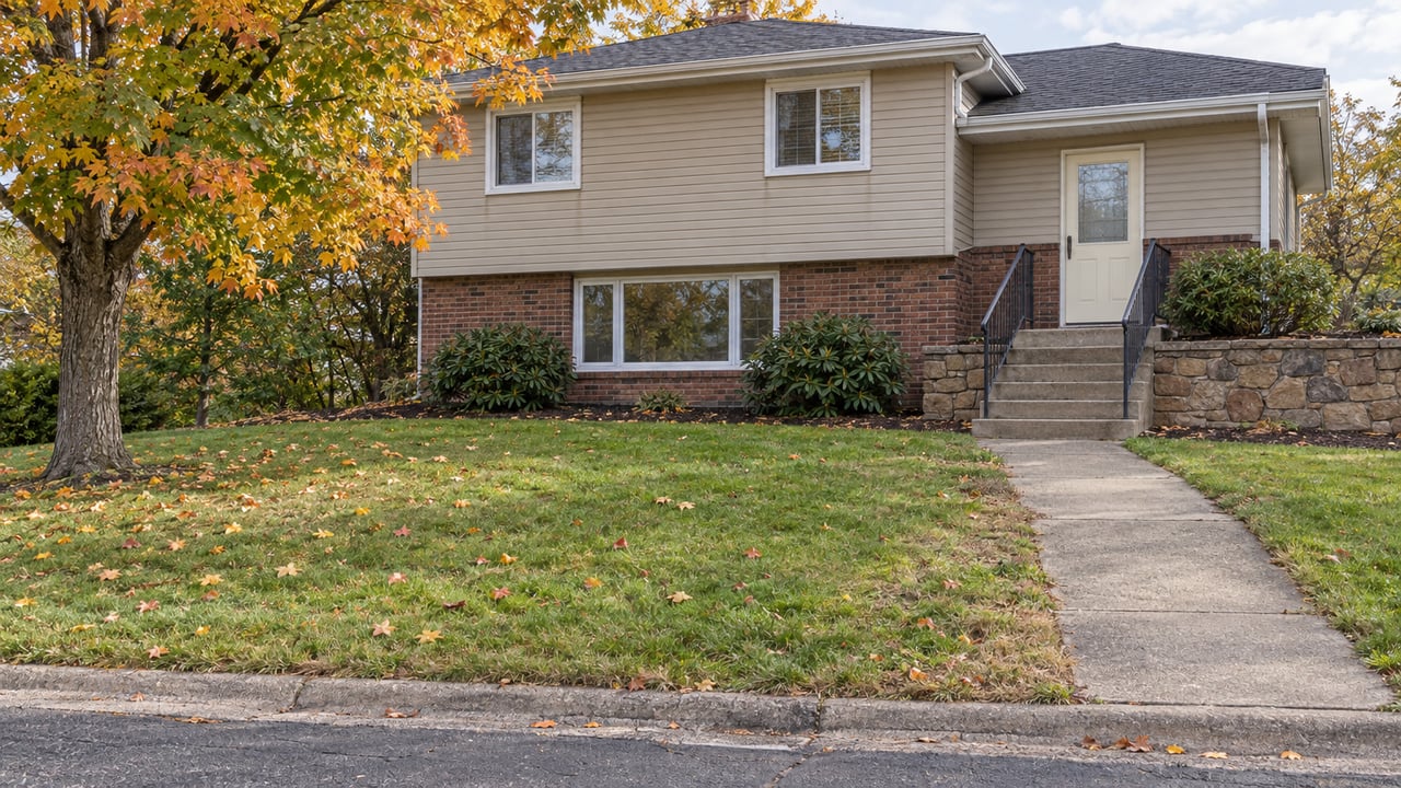 Brick and tan-sided split-level home above a sloping lawn, with concrete entry steps, sandstone retaining wall and a maple turning yellow and orange.