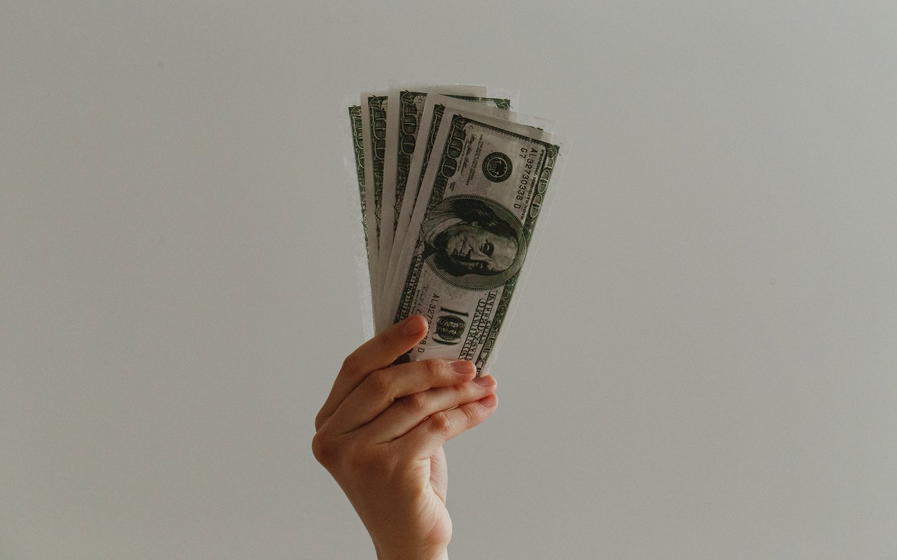 A hand holding a fan of U.S. hundred-dollar bills against a plain wall.