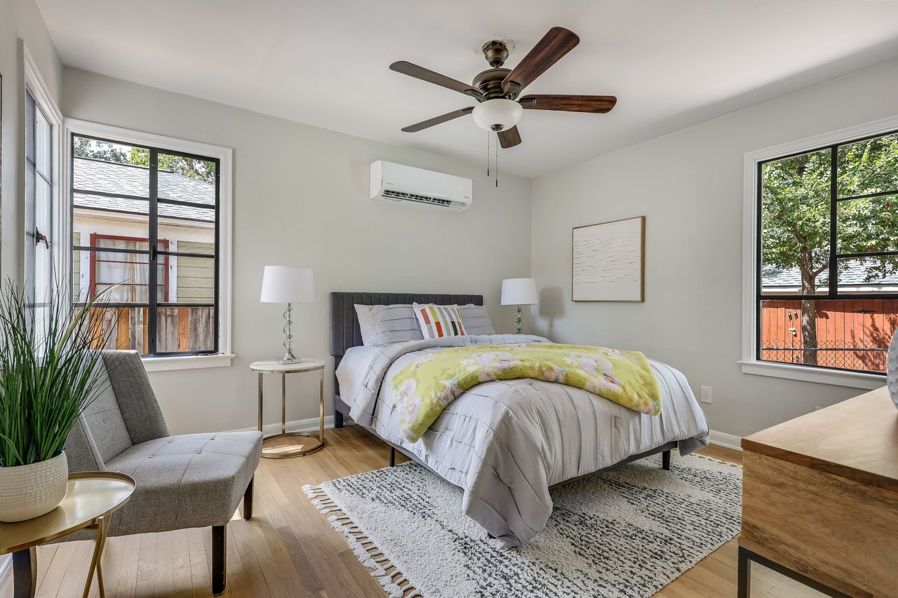 Bedroom with a white bed, pale yellow floral throw, gray chair, and large windows.