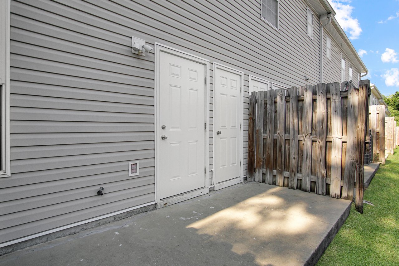 Gray siding of a house with two white doors; one opens to a fenced area. An outdoor light and grassy yard are present under a blue sky.