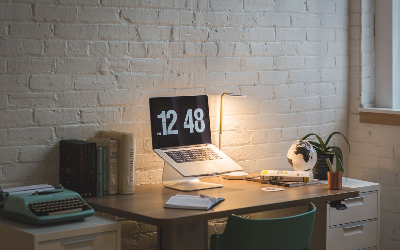 Home office with a laptop and lit desk lamp on a wooden desk against a white brick wall.