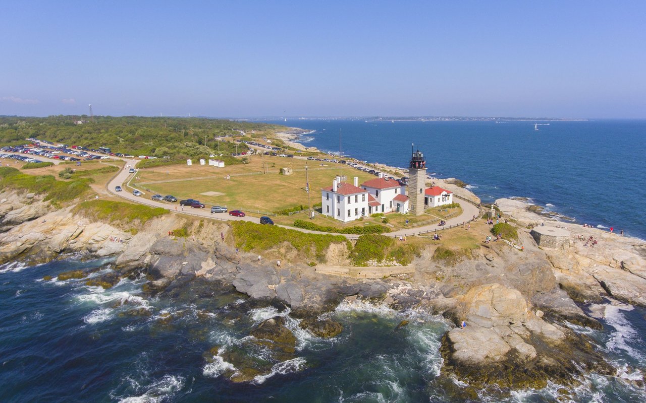 Beavertail Lighthouse on a rocky Jamestown shoreline, with white buildings, coastal road, and blue ocean.