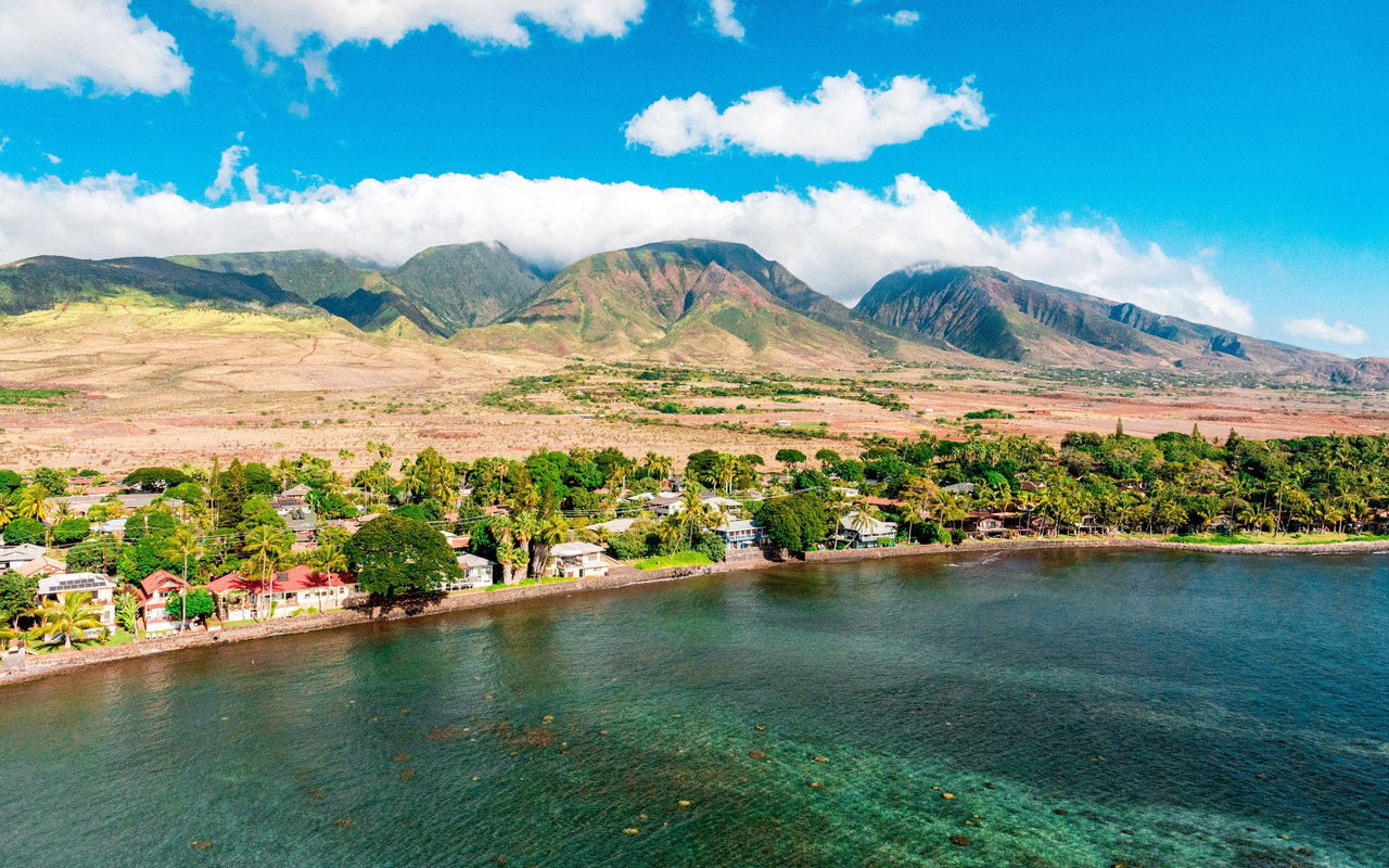 Aerial view of a coastal neighborhood beside clear water, with green mountains rising behind it.