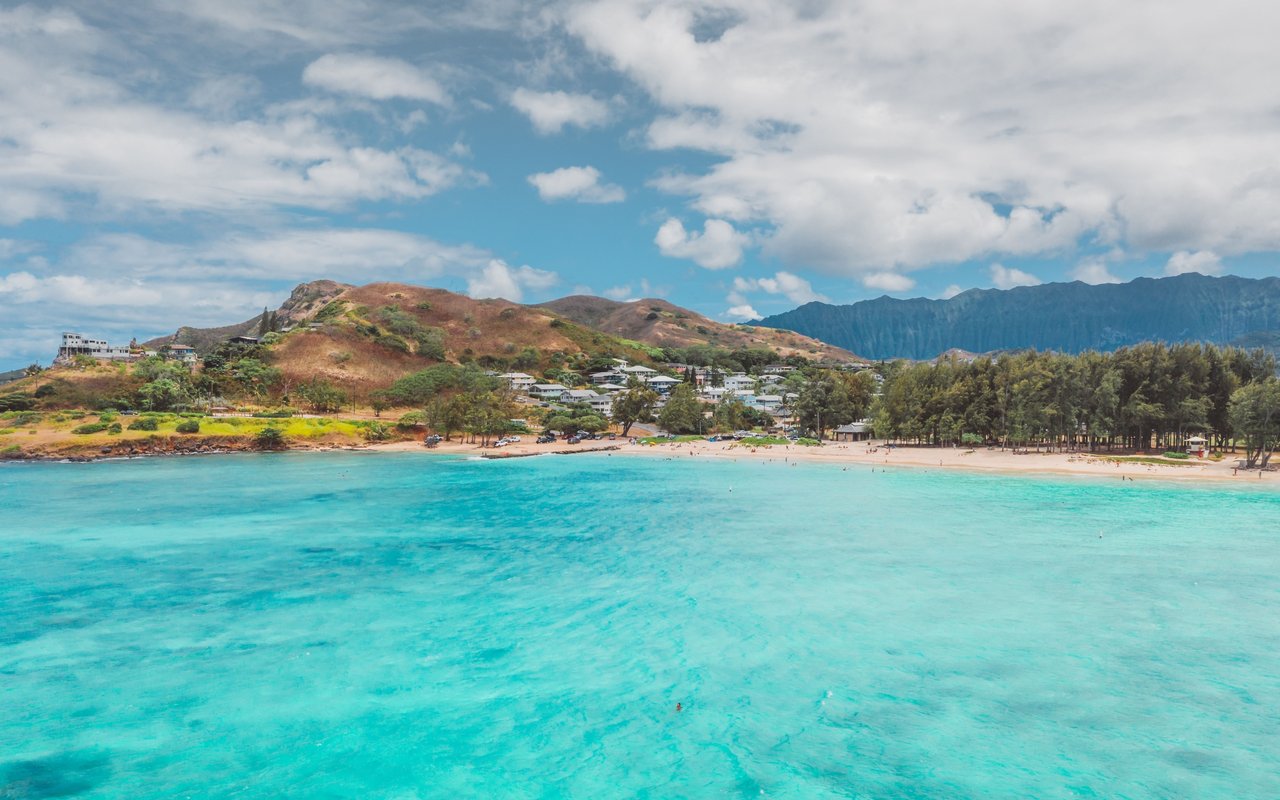 Turquoise bay and sandy beach below hillside homes and a rugged mountain ridge.