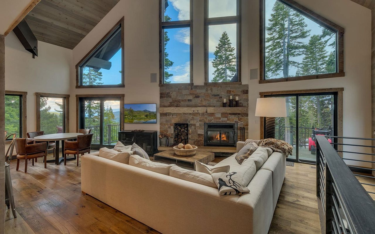 Furnished living room with a stone fireplace, vaulted wood ceiling, and tall windows overlooking pine trees.