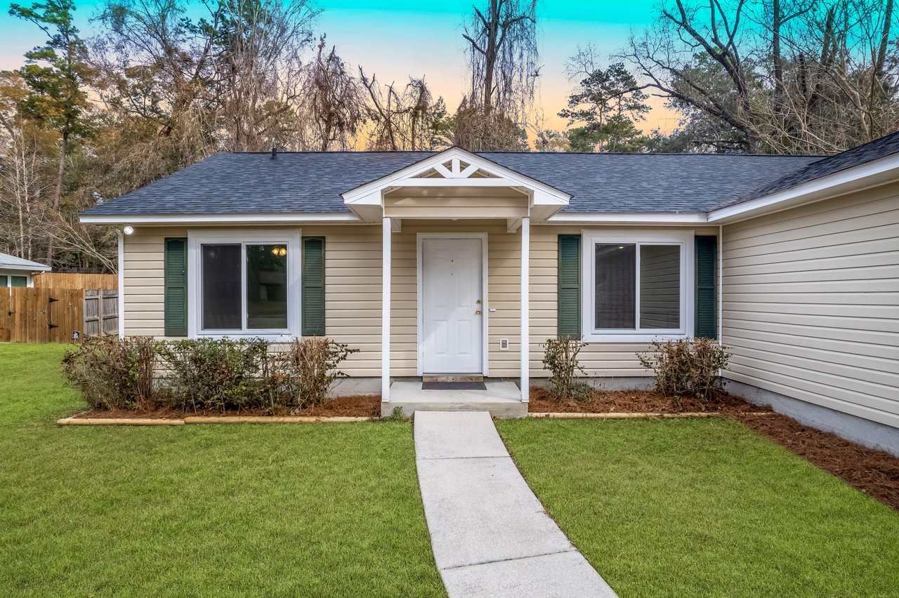 A cozy single-story house with beige siding and white trim. A concrete pathway leads to a covered entrance. The yard is neatly landscaped.