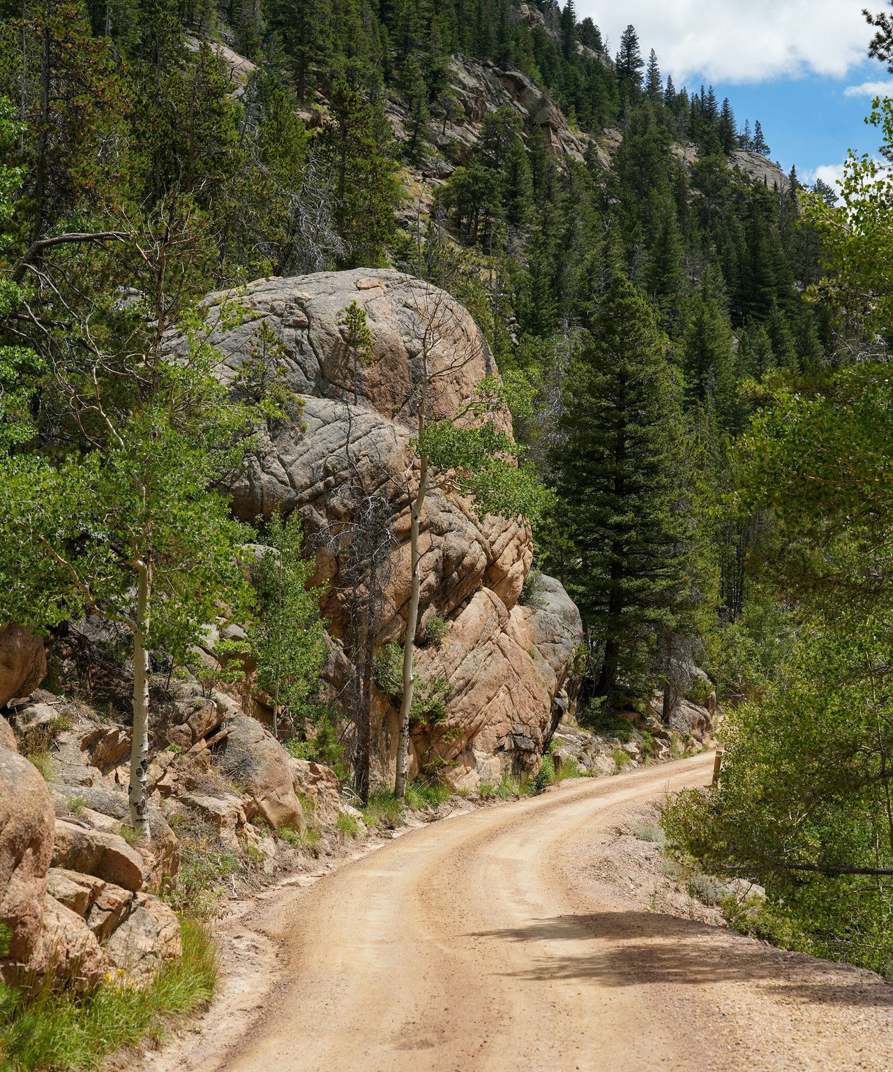 Dirt road curving past granite boulders and evergreen trees on a sunny mountain slope.