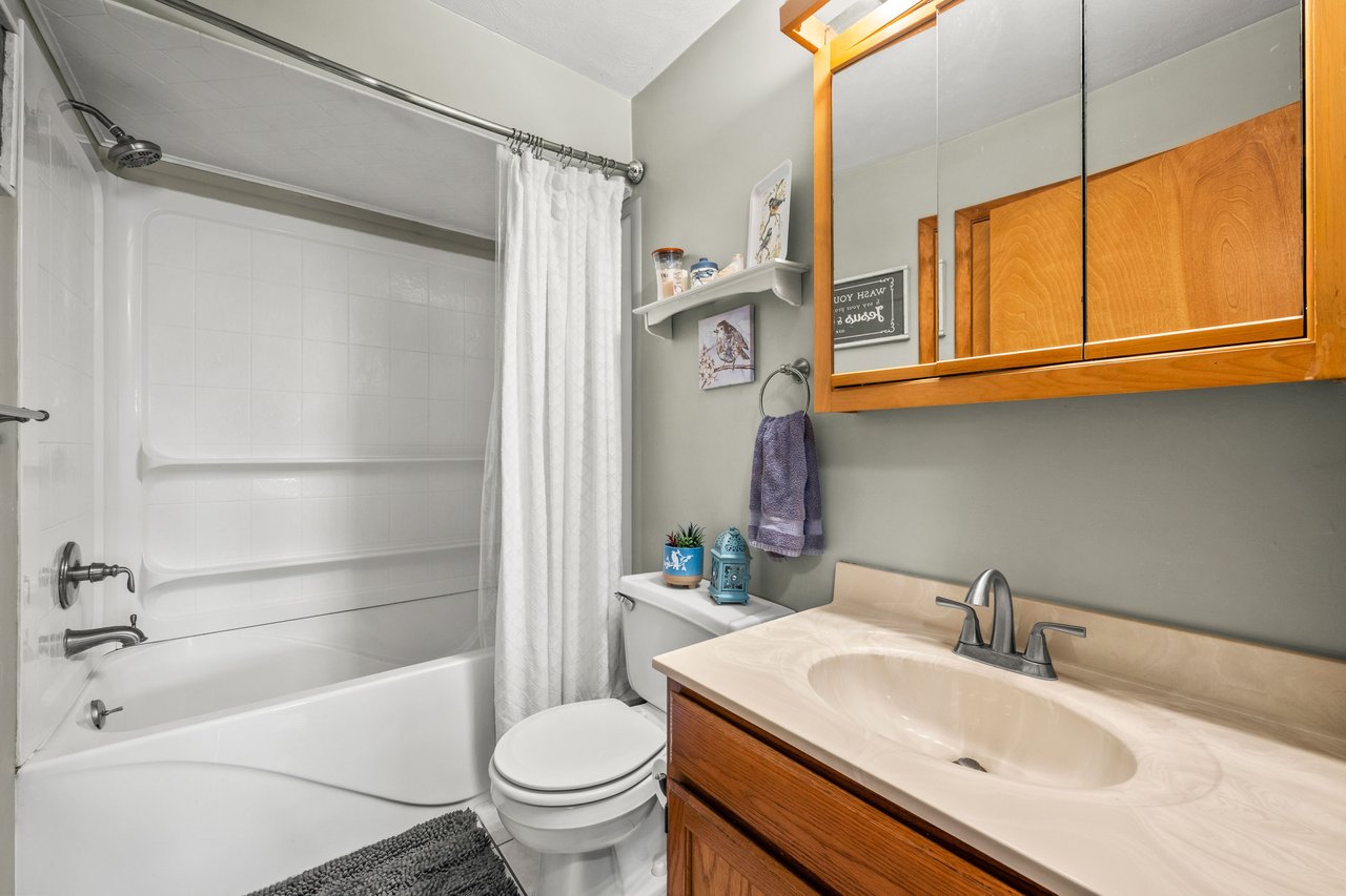 Bathroom with a white tub and shower, toilet, and cream vanity beneath mirrored wood cabinets.