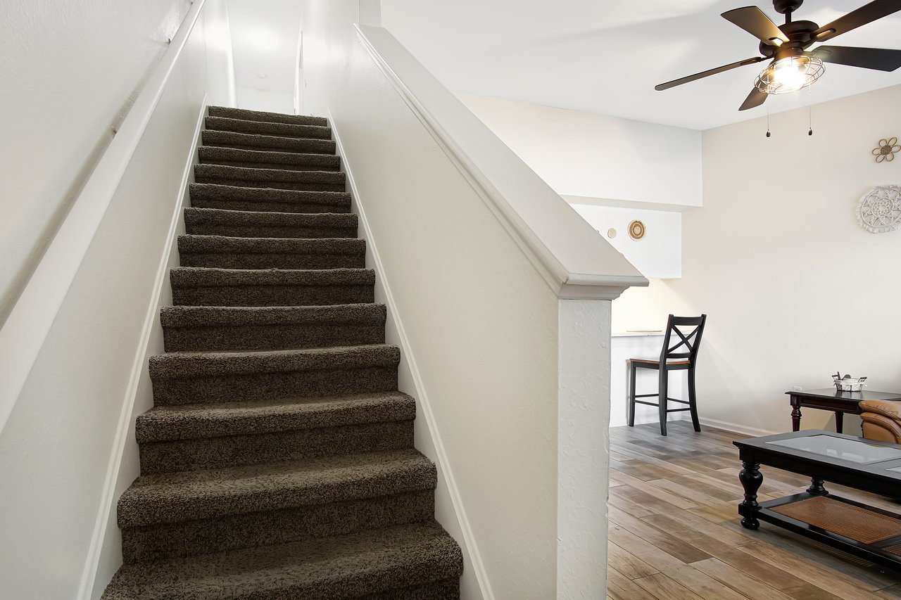 Carpeted staircase with white walls leads to upper floor. Right side shows a modern room with tiled floor, black chair, and ceiling fan. Bright and airy.