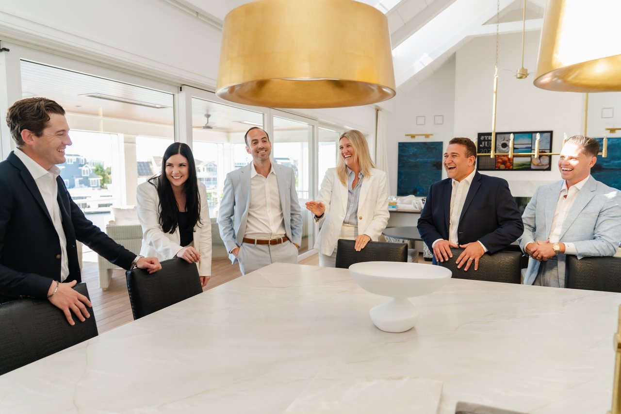 Six real estate agents smiling around a conference table in a bright, modern office.