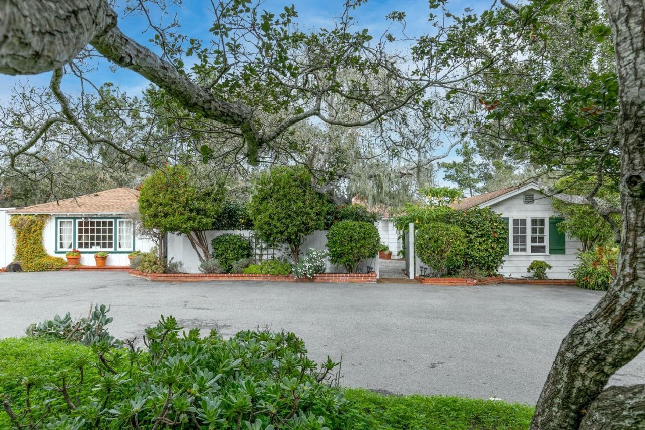 White house with green-trimmed windows, framed by mature trees and landscaped garden beds.