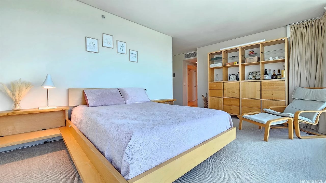 Furnished bedroom with a large bed, built-in wooden shelving, and a cushioned chair by curtained windows.