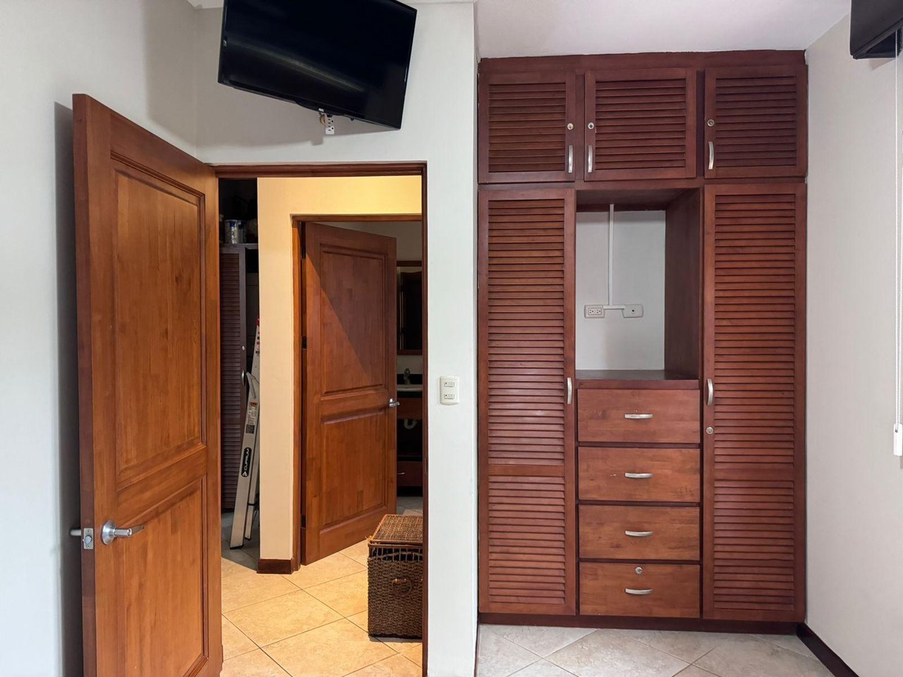 Bedroom with a built-in wooden wardrobe, drawers, wall-mounted television, and open doorway.