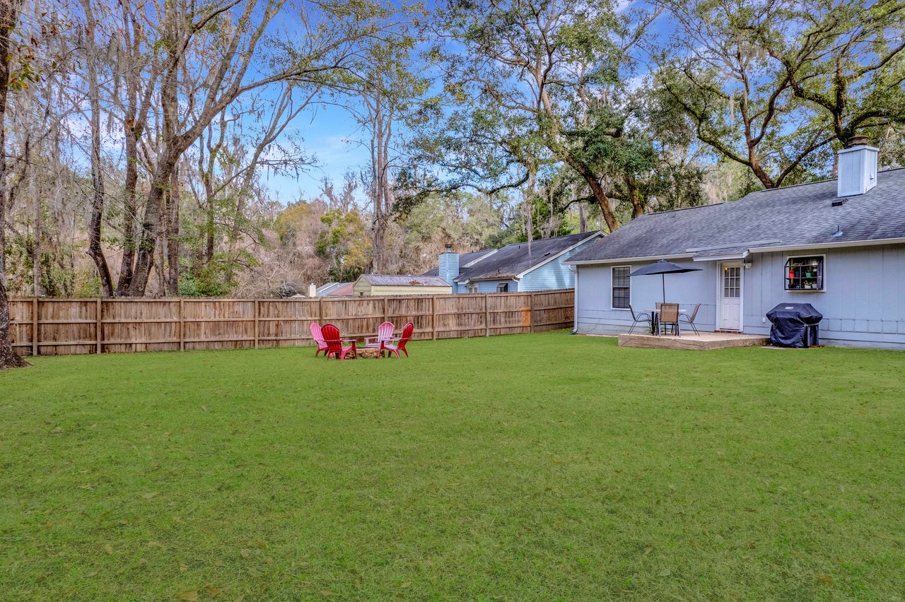 A spacious backyard features vibrant green grass, a cluster of four red Adirondack chairs around a fire pit. The blue house has a patio with a table, chairs, and a barbecue grill. Trees and a wooden fence surround the yard under a clear blue sky.