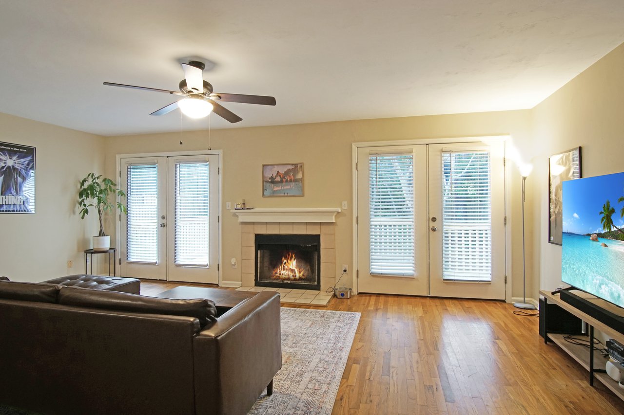 Living room at 529 Collinsford Road, Tallahassee, Florida, featuring a cozy fireplace with tile surround, hardwood floors, and two sets of French doors with blinds that open to the back deck. The space includes a brown sectional sofa, area rug, and a ceiling fan with light fixture, creating a bright and inviting atmosphere.