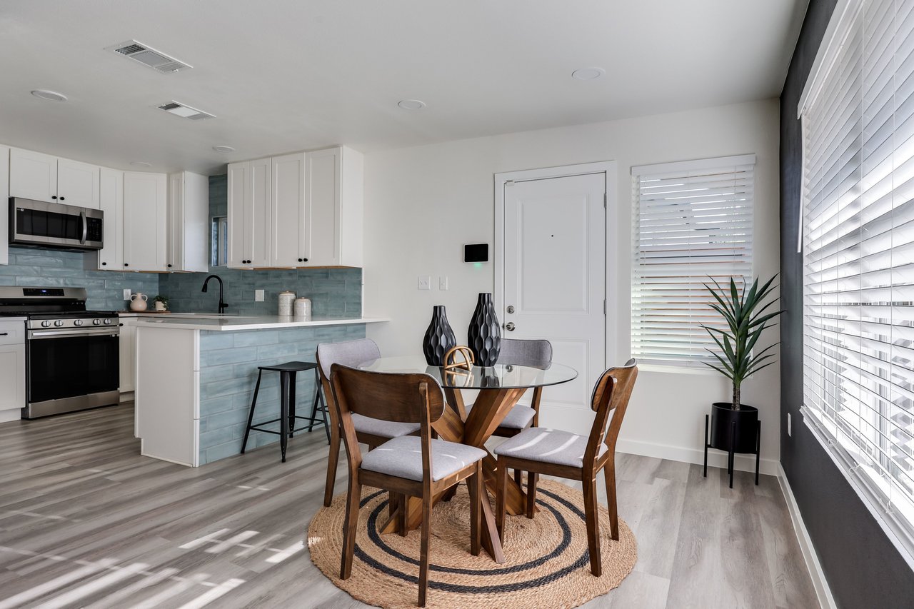 Open-plan kitchen and dining area with blue-gray tile backsplash and a round table beneath large windows.