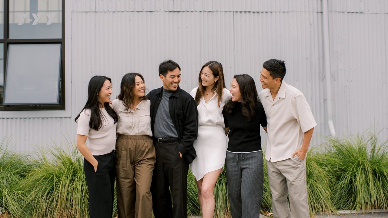 Six smiling real estate team members stand together outside a corrugated building.