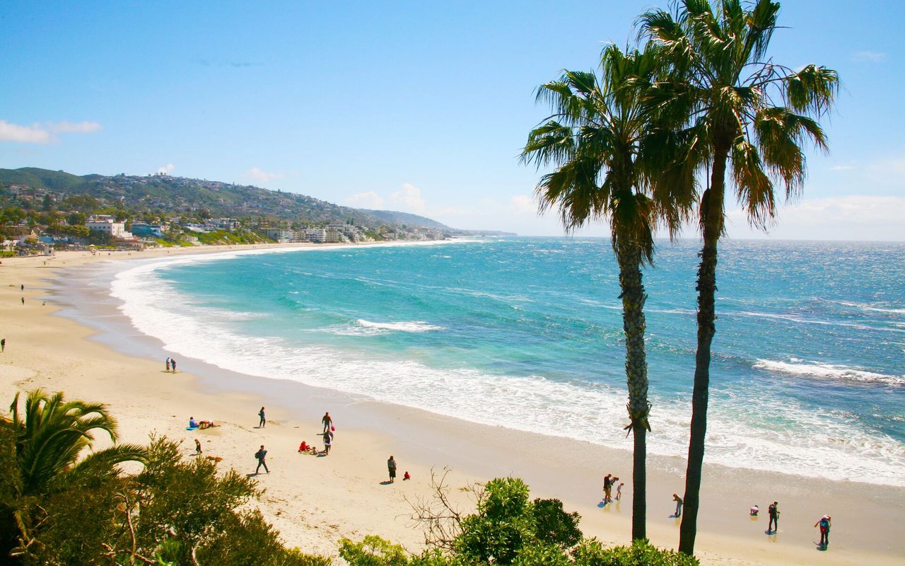 Sandy beach and turquoise coastline framed by palm trees, with hills and buildings along the shore.