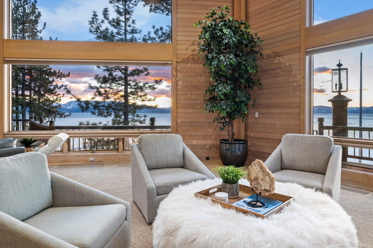 Living room with tall windows overlooking a lake and snow-dusted mountains at sunset.