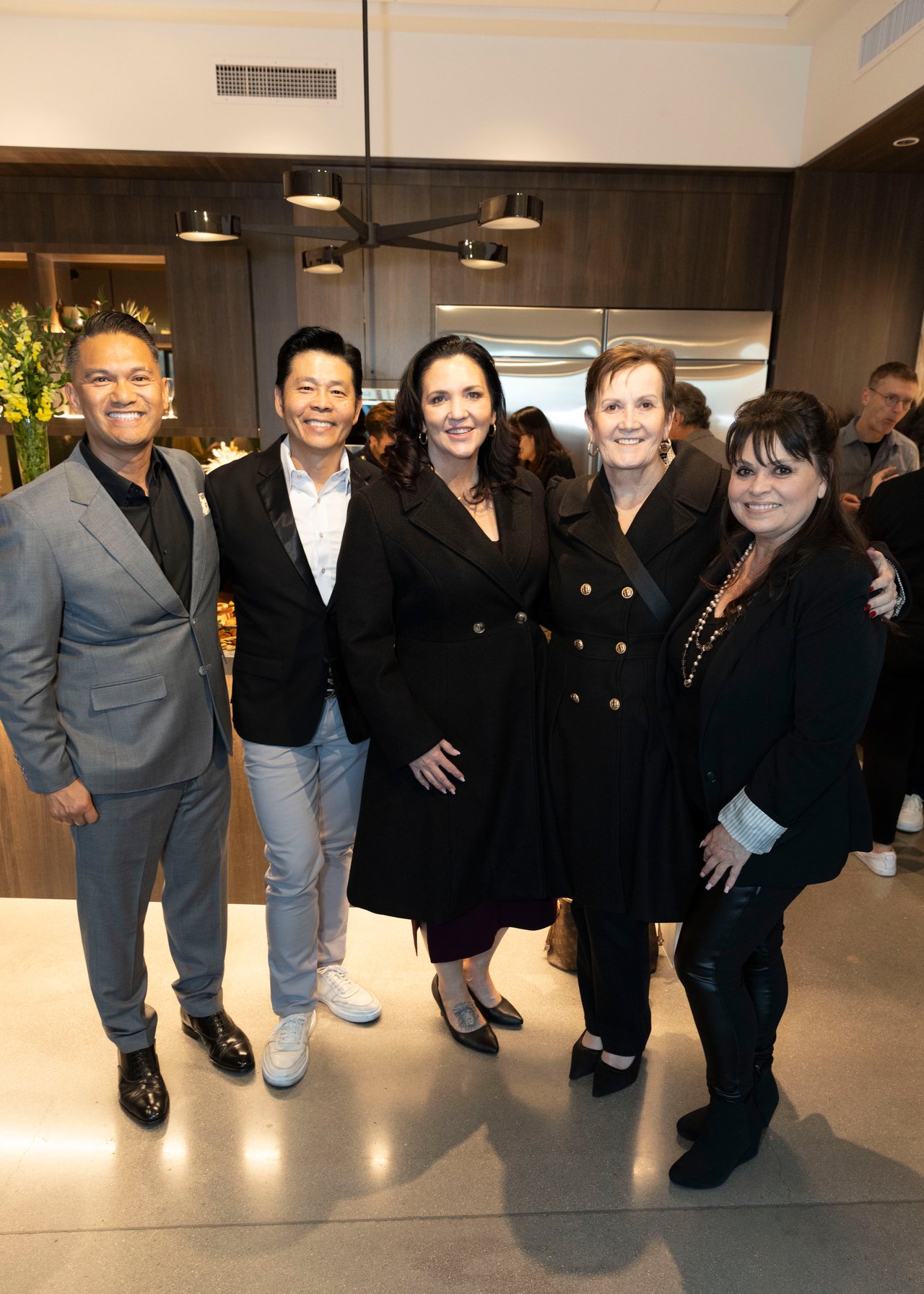 Five attendees pose together at an indoor open house, with guests gathered behind them.