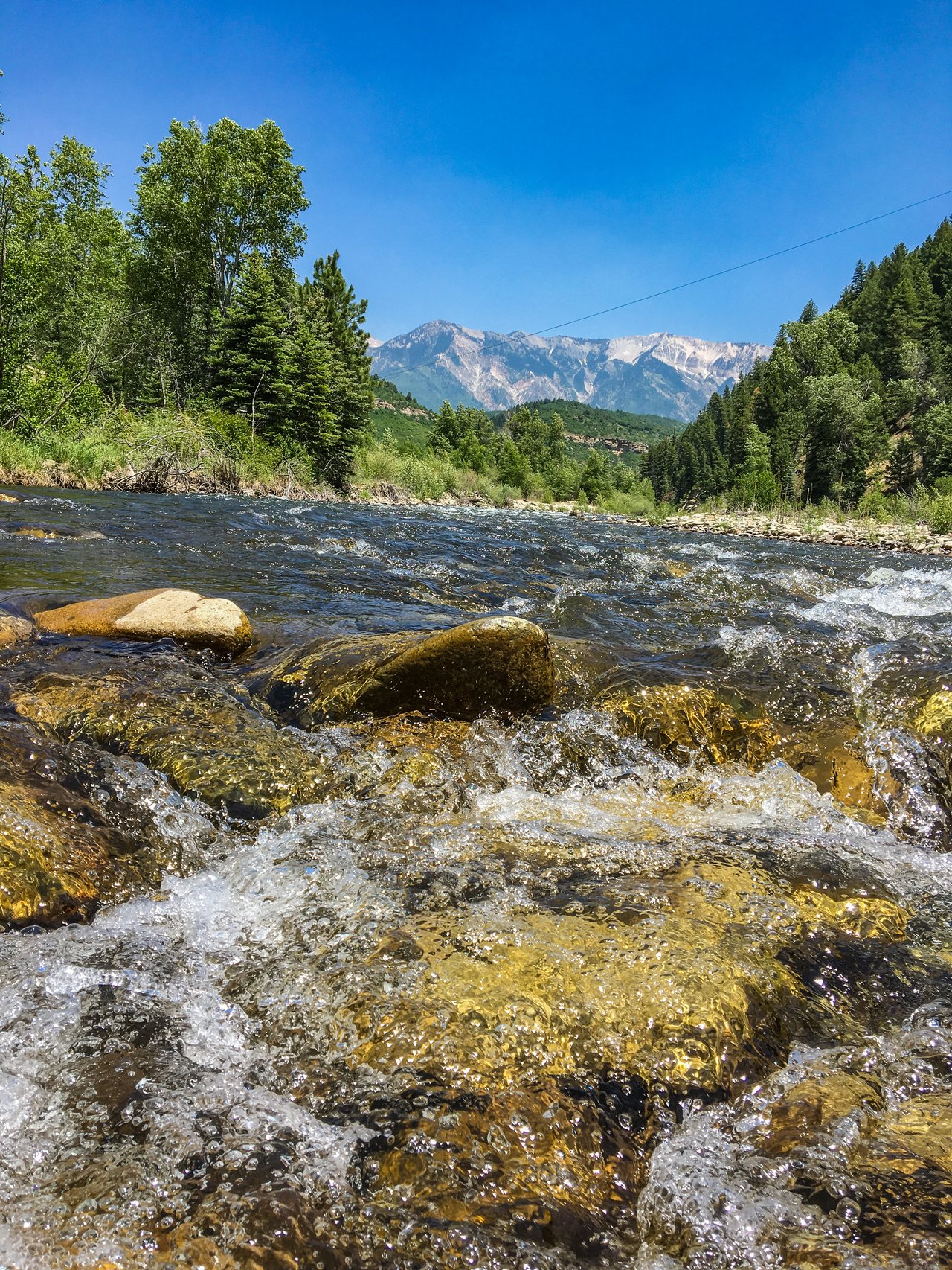 Clear river flows over rocky shallows beneath forested hills and rugged mountains.
