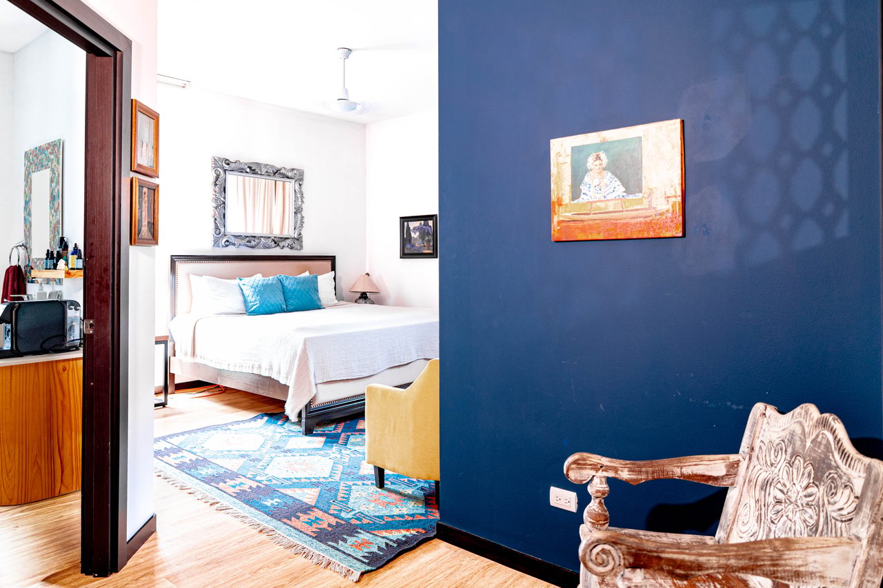 Furnished bedroom with a white bed, turquoise pillows, colorful rug, and deep blue accent wall.