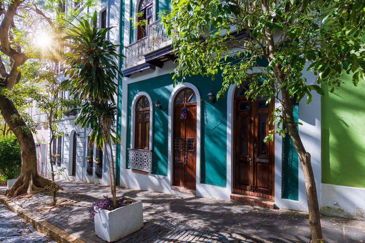 Teal and white building with arched wooden doors, ornate balcony, and leafy trees.