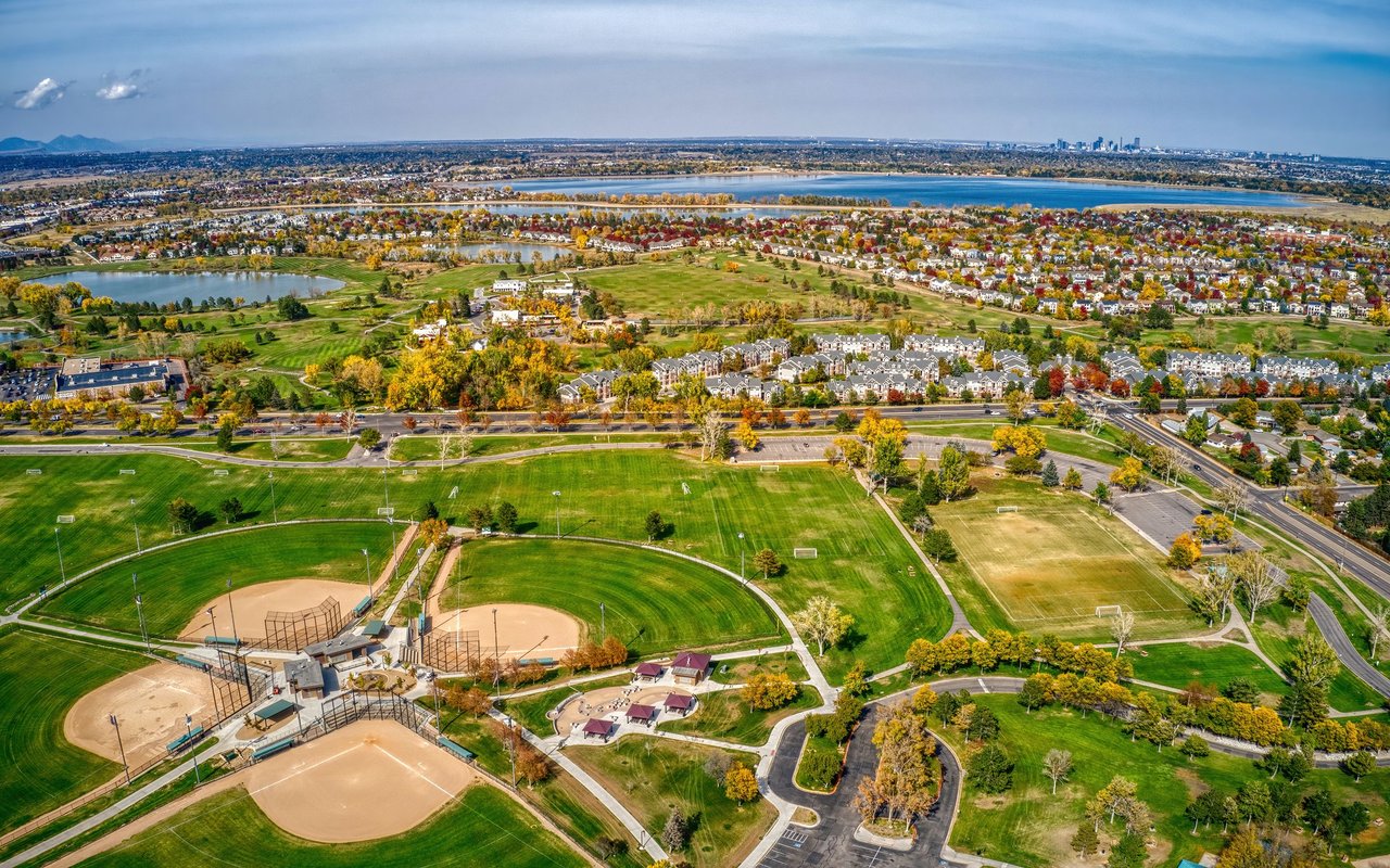 Aerial view of suburban homes and parks, with baseball fields, water, and mountains in the distance.