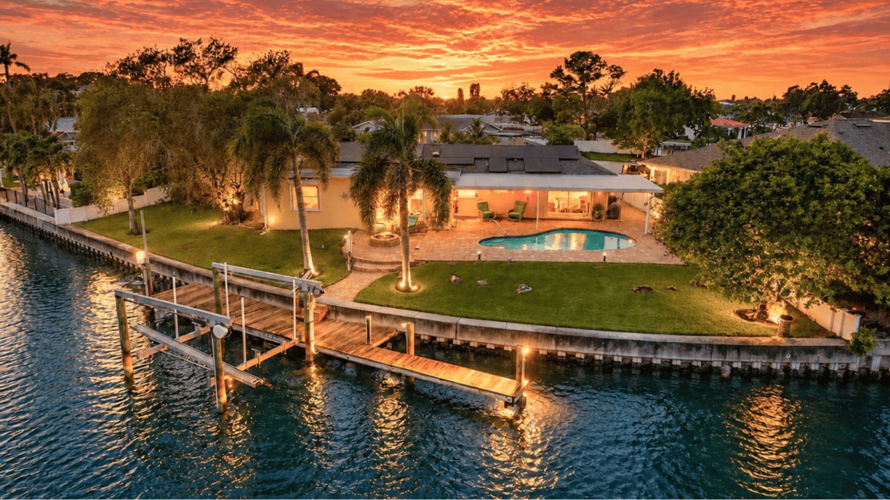 Waterfront home with a pool and dock beside a canal under an orange sunset.