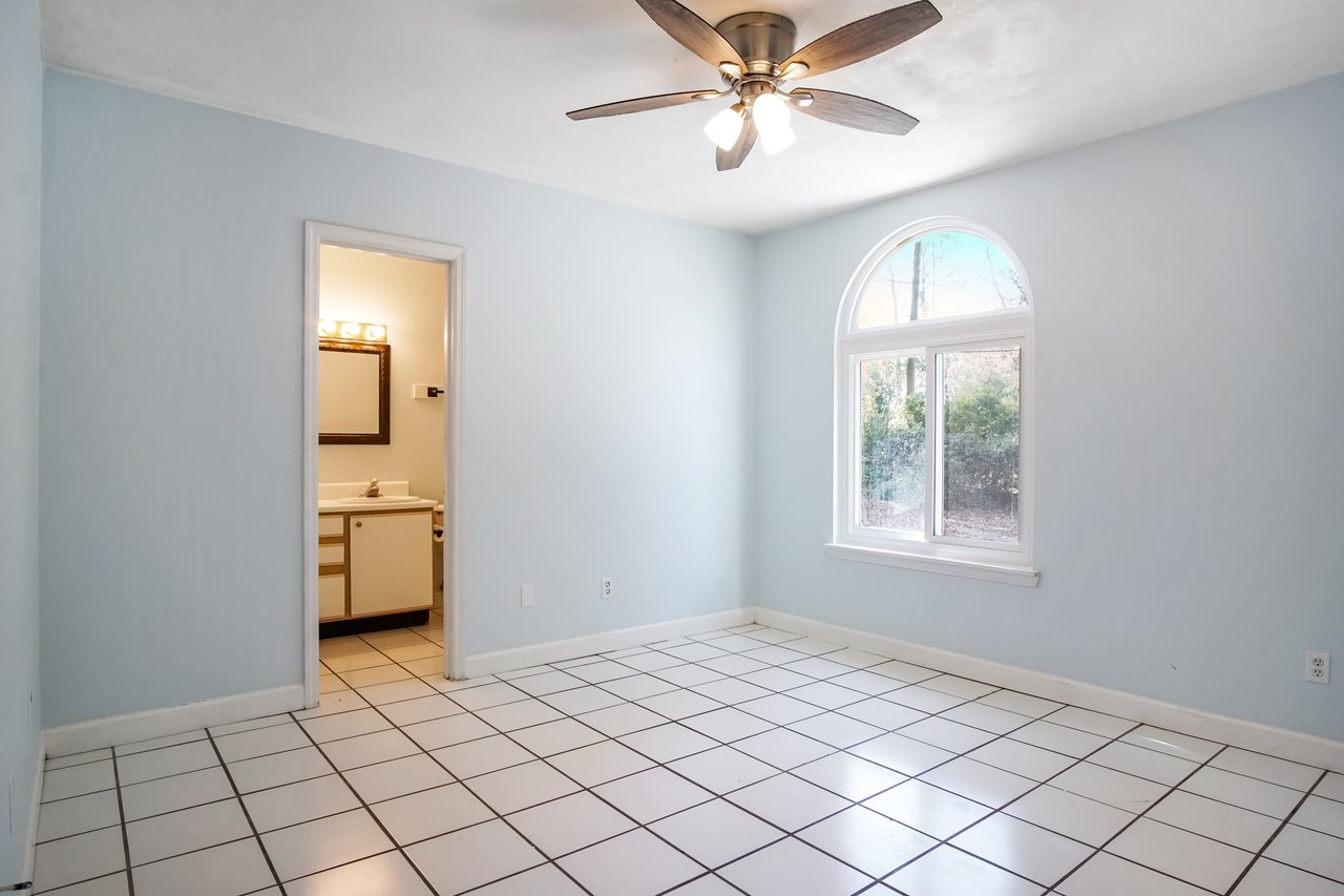 Empty room with light blue walls and white tiled floor, featuring an arched window showing greenery outside. A ceiling fan is centered above, and a lit bathroom with a large mirror is visible through an open door.