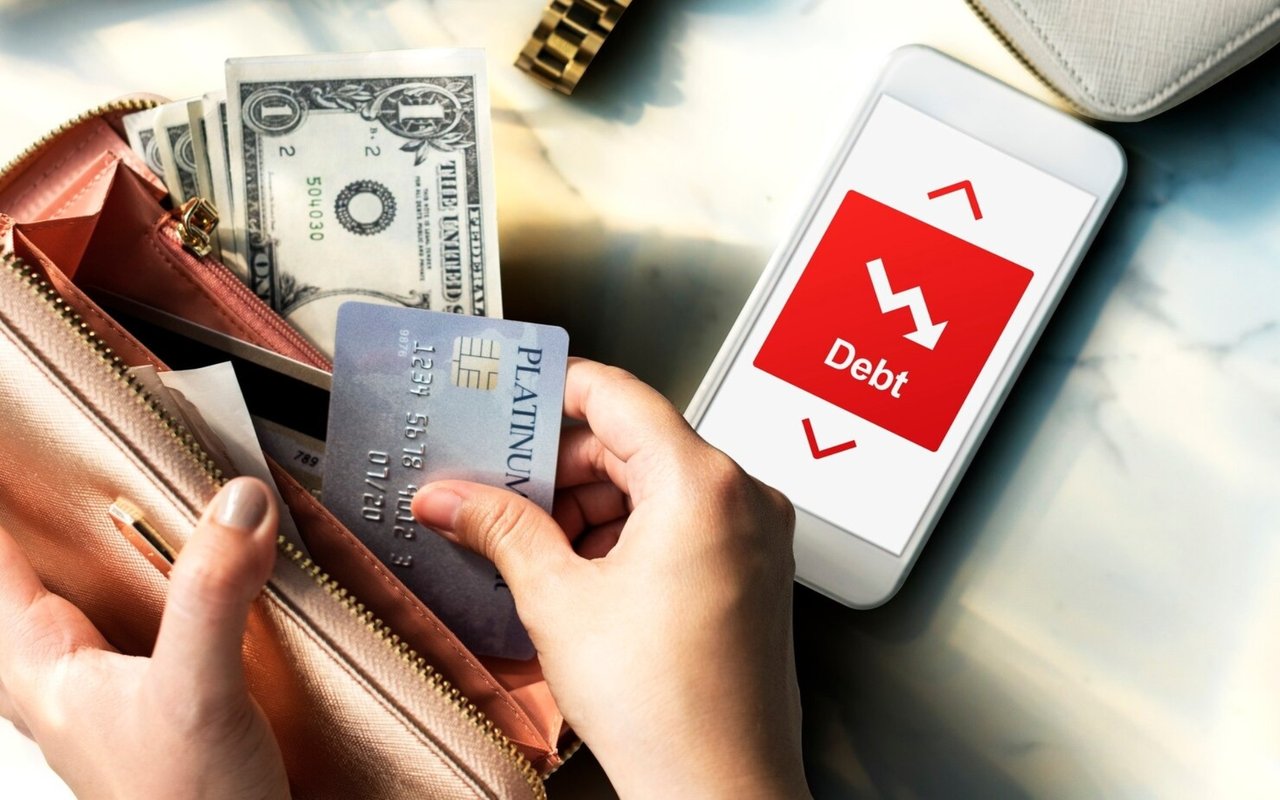 Hands holding a platinum credit card over a cash-filled wallet beside a phone displaying “Debt.”