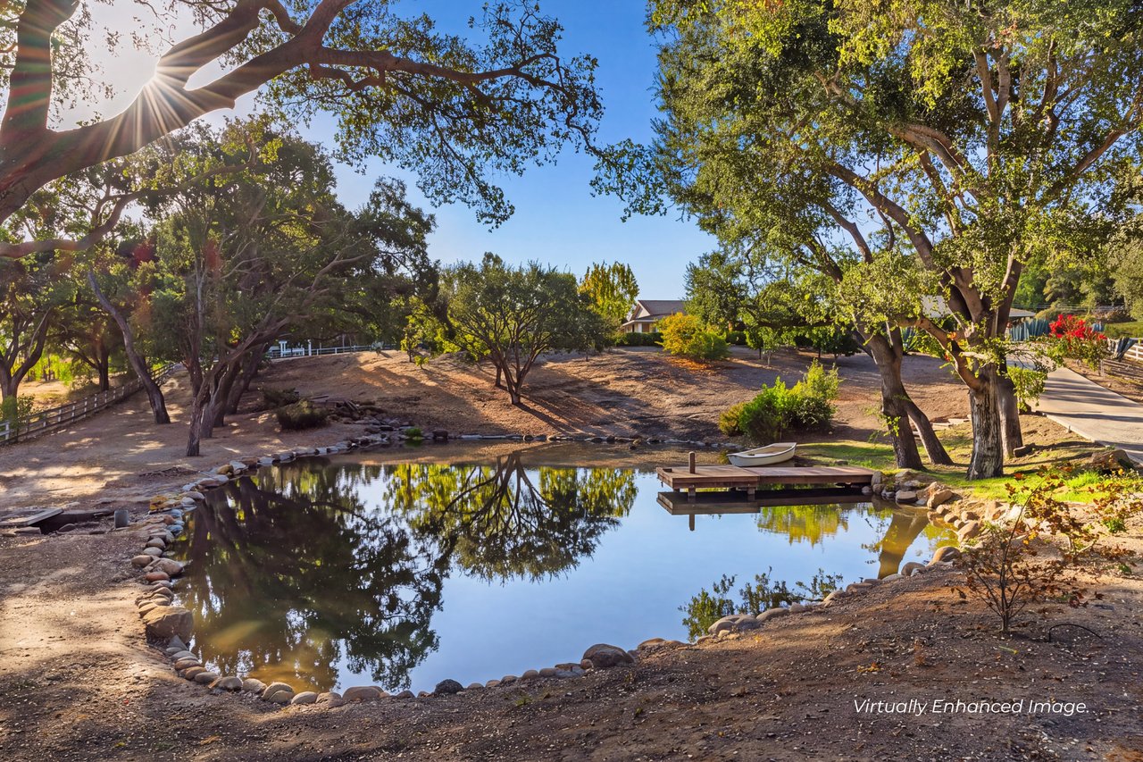 Property grounds with a tree-lined pond, small dock, boat, and distant home.