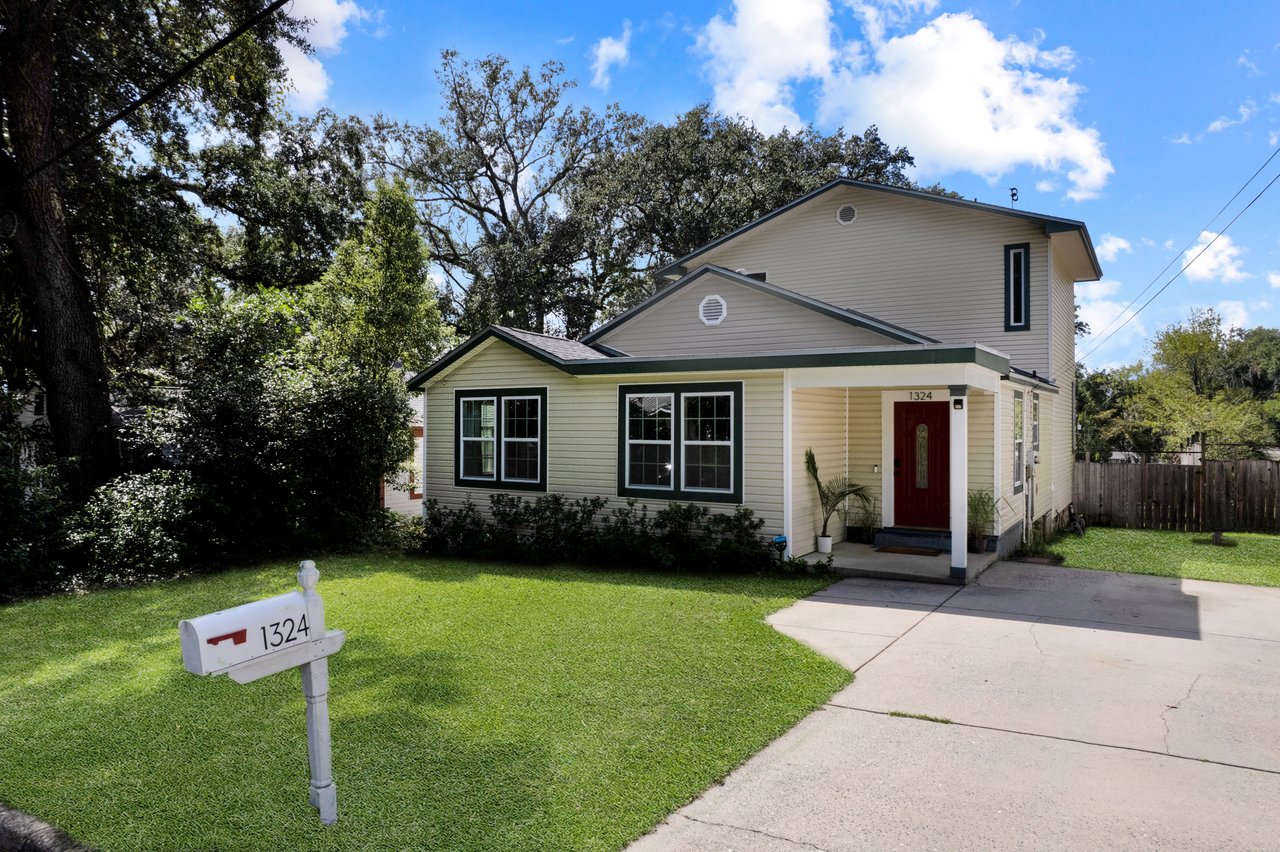 A charming, two-story white house with green trim, featuring a red door, is surrounded by lush greenery and a well-manicured lawn under a blue sky at 1324 Branch Street in Tallahassee, Florida.