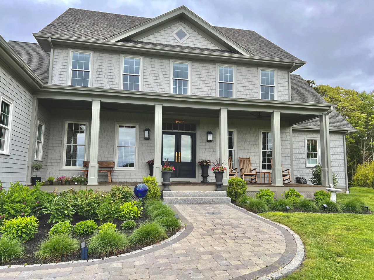 Gray-shingled two-story house with a covered porch, black double doors, and a curved stone walkway.