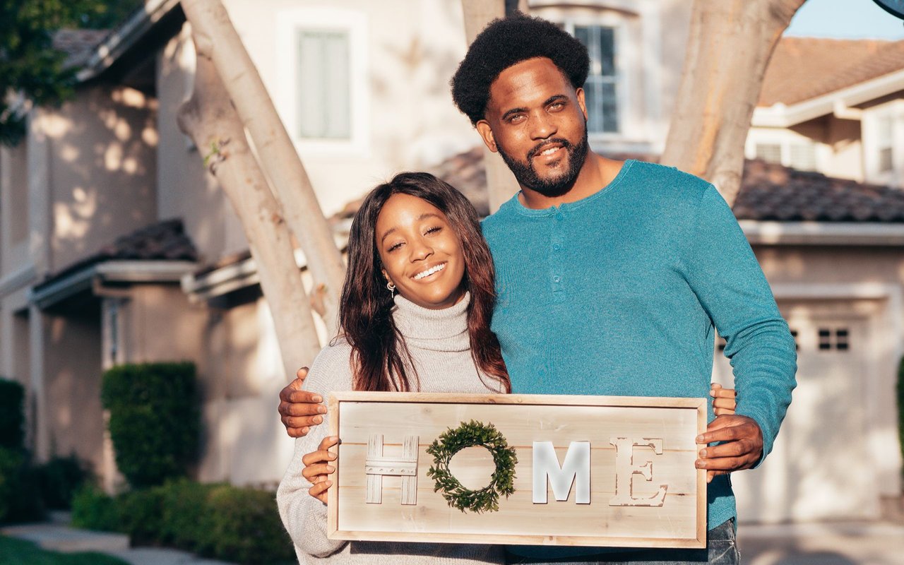 Smiling couple outside a house holding a wooden HOME sign.