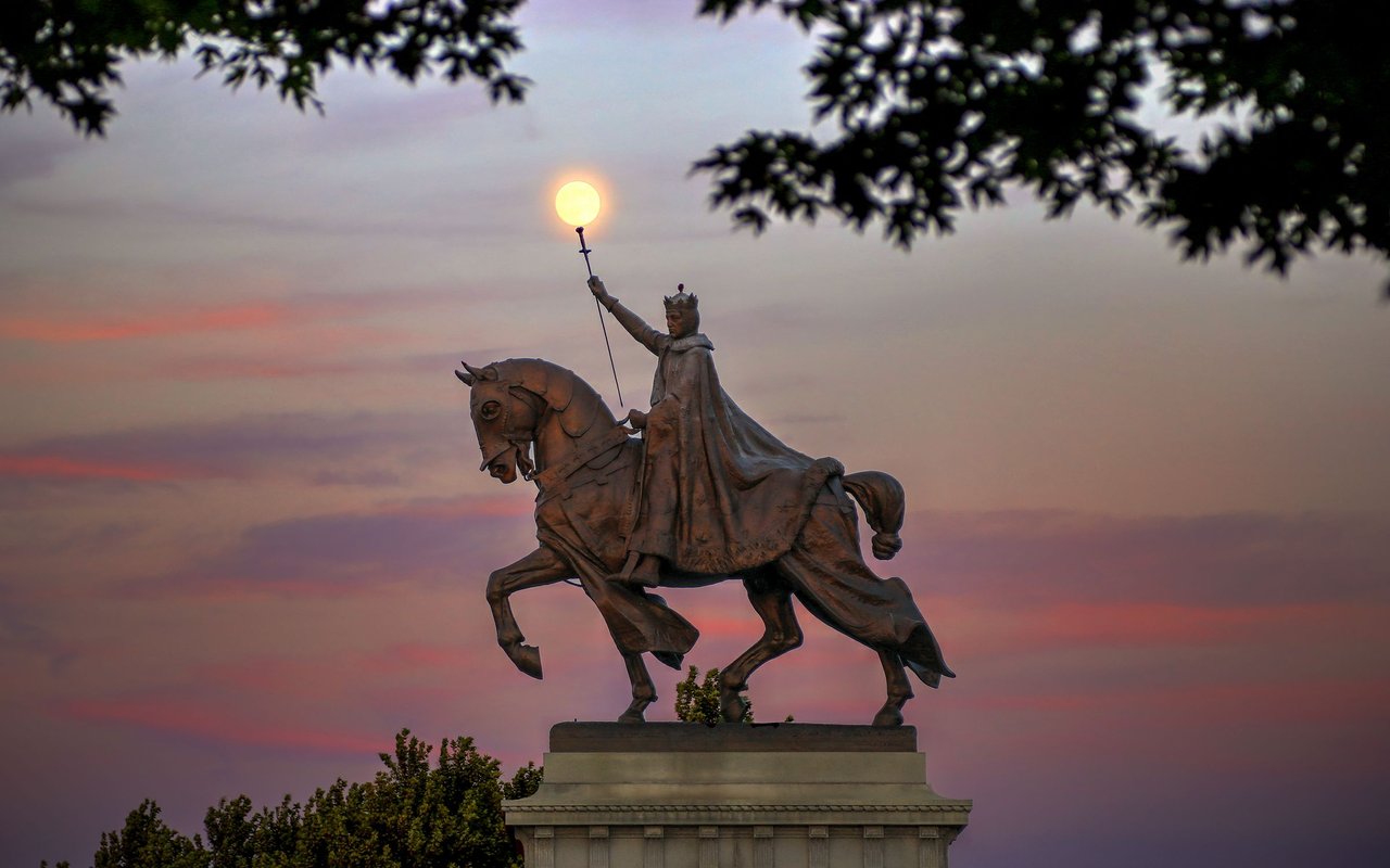 Equestrian statue of Saint Louis against a pink sunset, framed by tree branches.