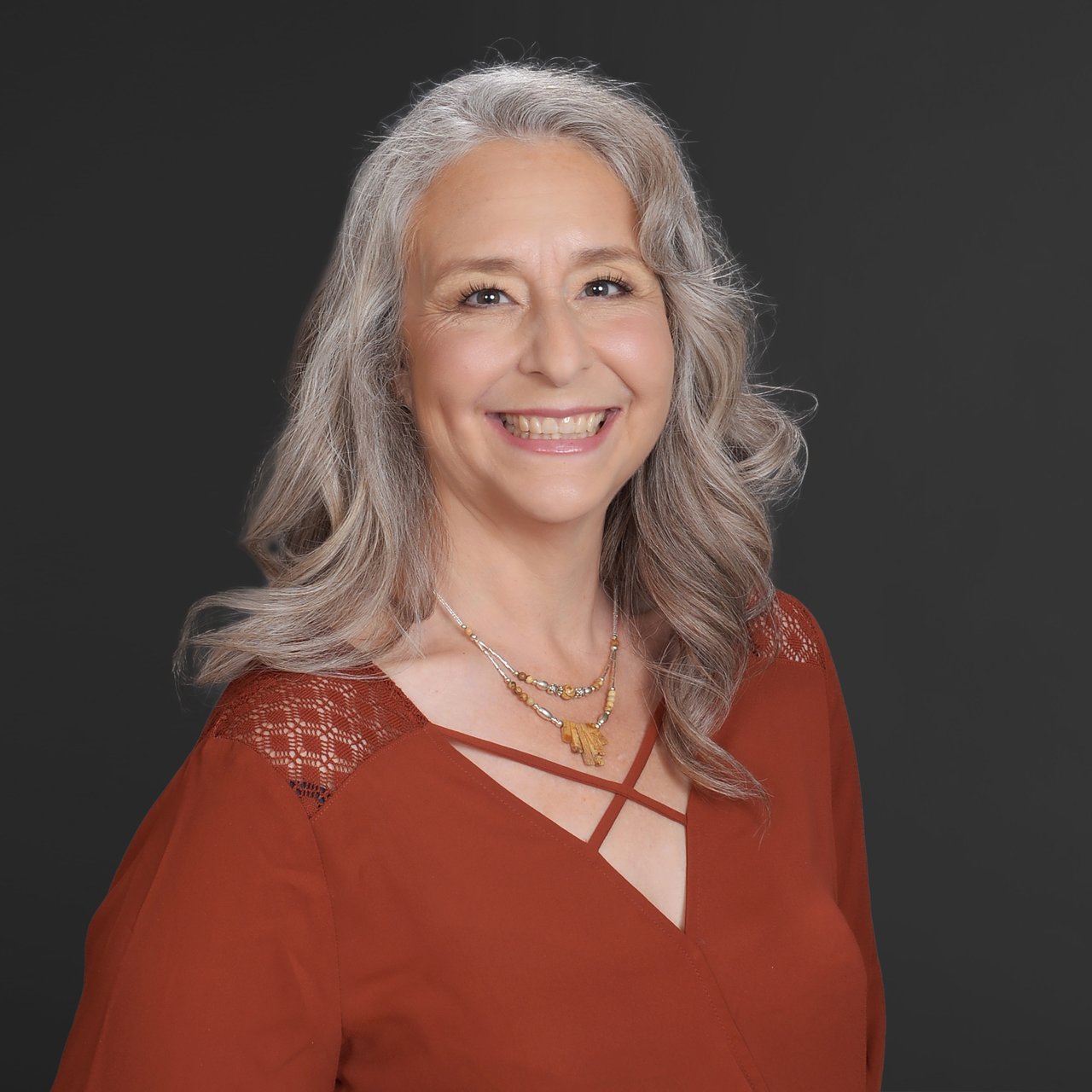 Elizabeth Xydas smiling in a rust-red blouse against a dark background.