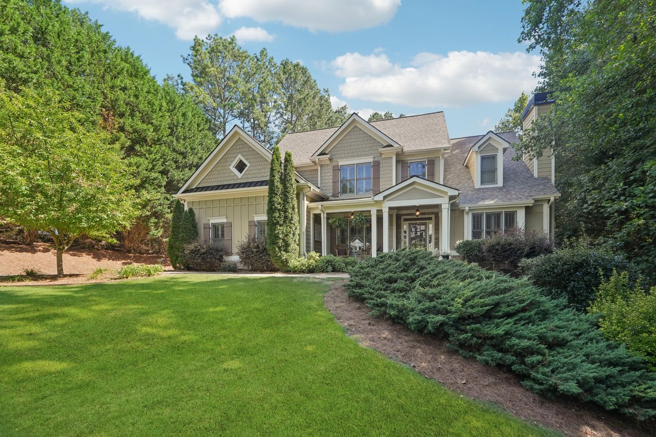 Two-story beige house with a covered porch, mature trees, and a broad green lawn.