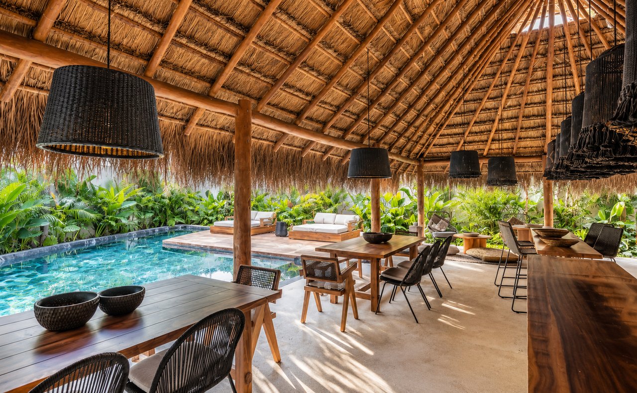 Thatched dining pavilion beside a pool, furnished with wooden tables and woven chairs amid tropical greenery.