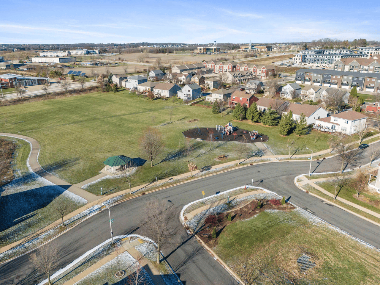 An aerial drone view of a quiet Verona, WI neighborhood featuring a central park, playground, and walking paths, highlighting the family-friendly "Hometown USA" lifestyle in 2026.