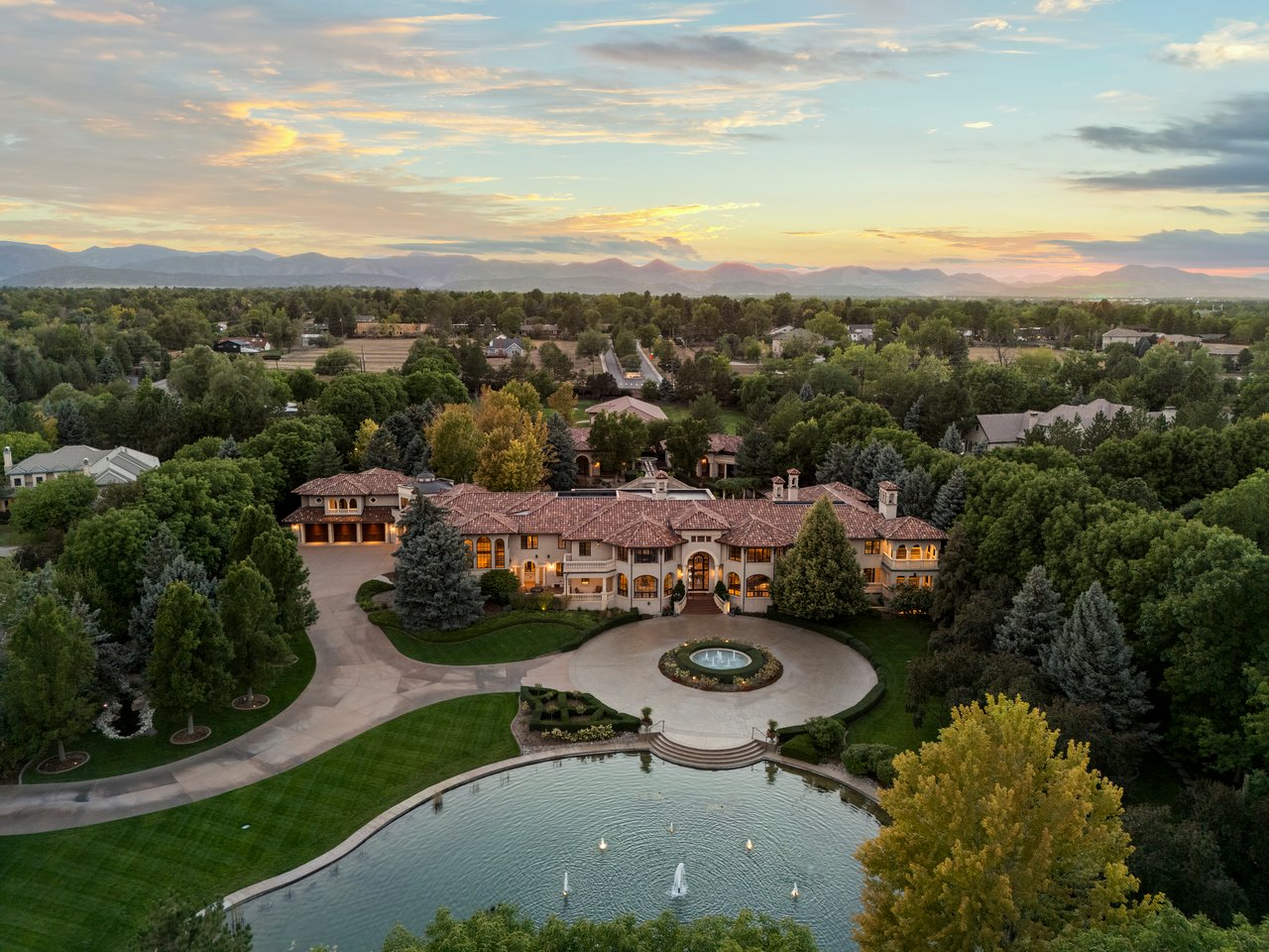 Aerial view of a grand estate with a circular driveway, fountain, swimming pool, and mountains at sunset.