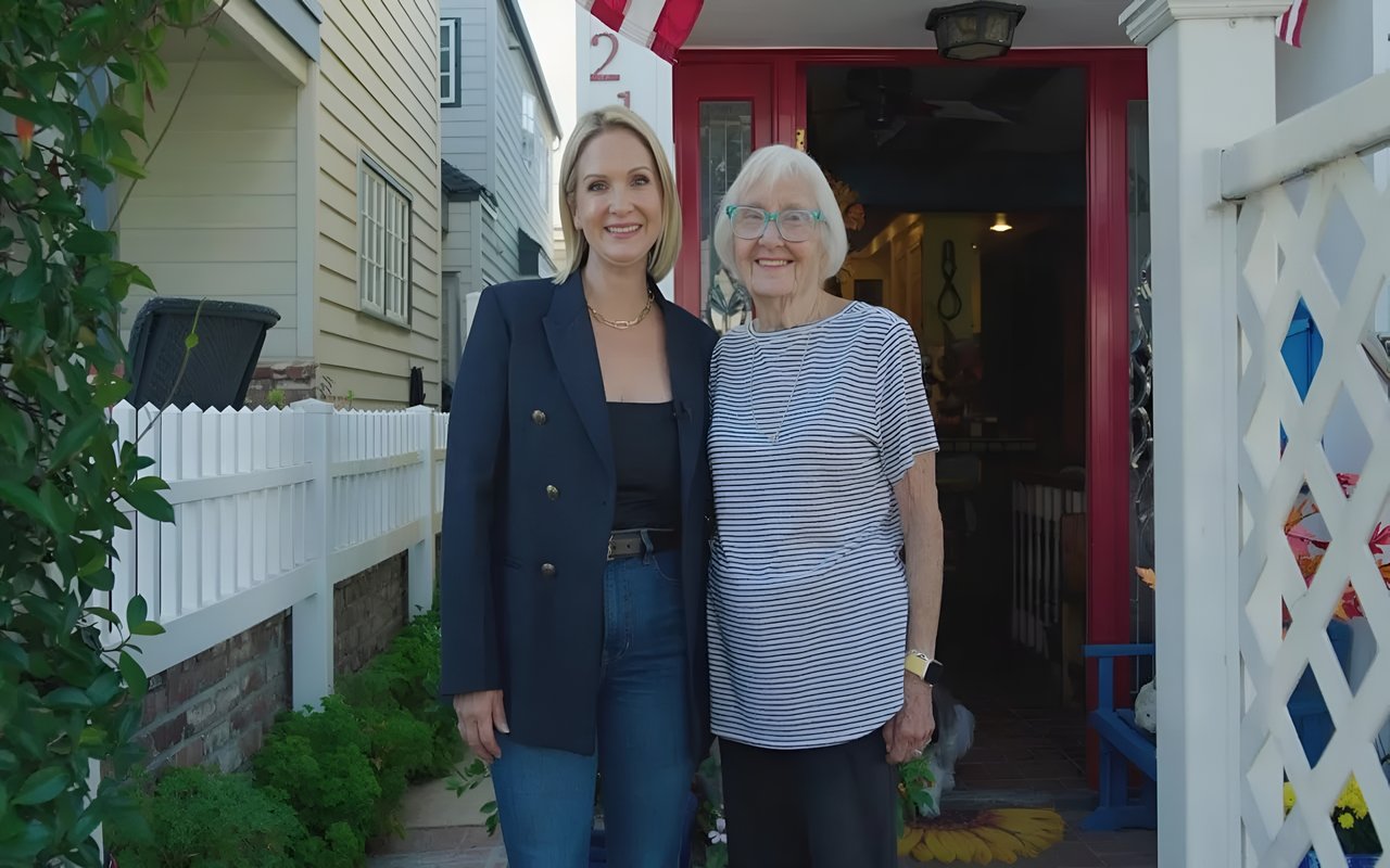 Annie Clougherty and an older woman stand in front of a red door, posing for a photo. A white picket fence is to their left.