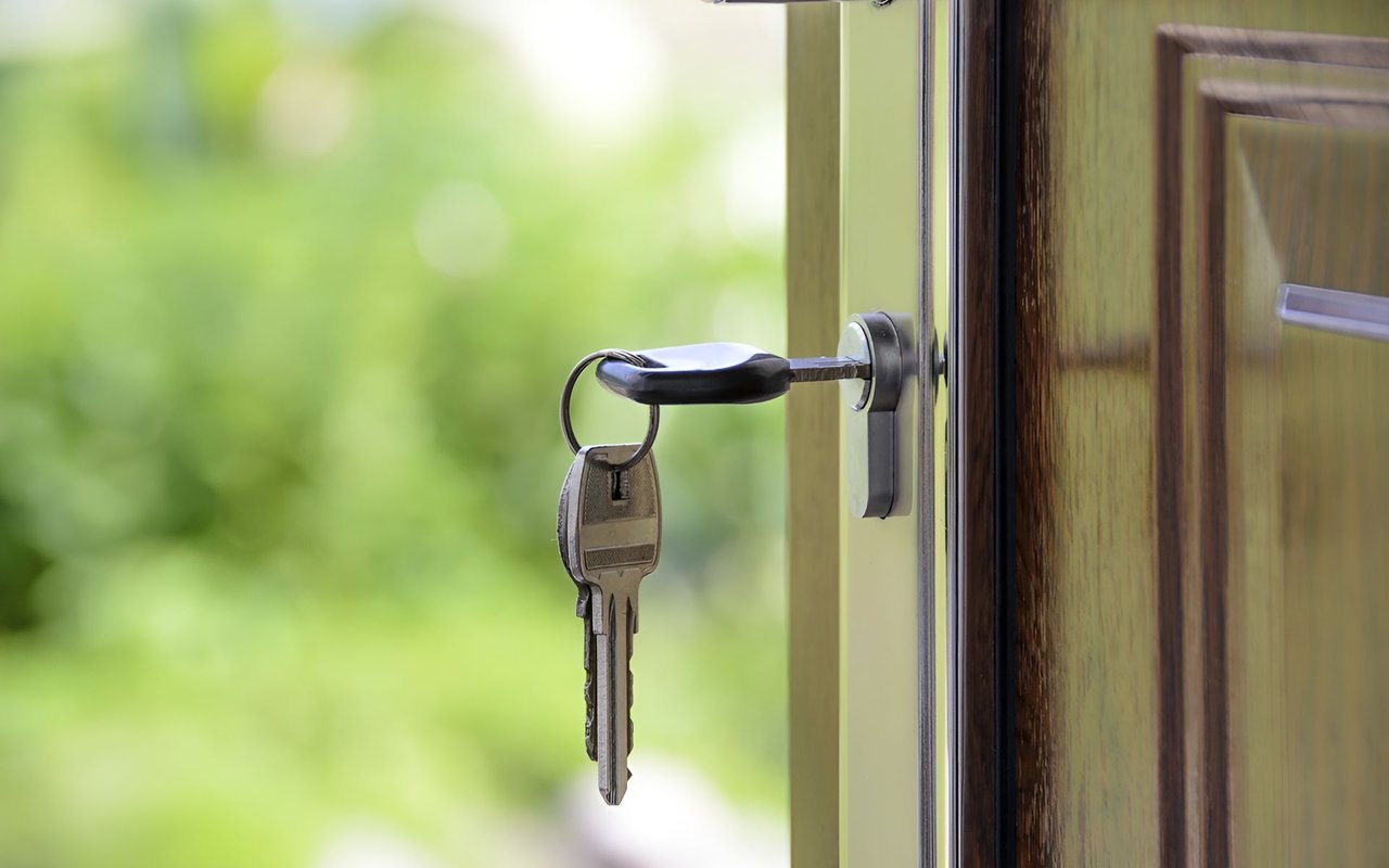 Key in the lock of a wooden front door, with green foliage outside.