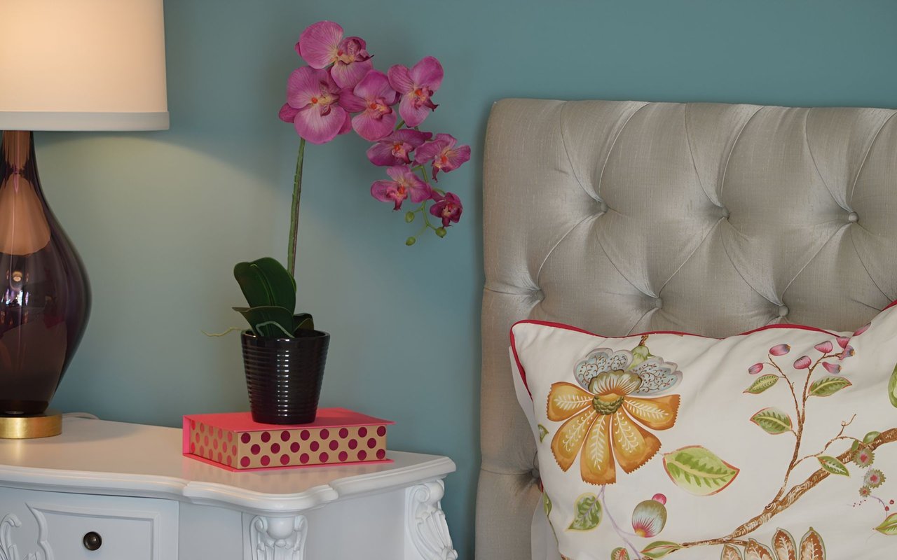 Bedroom with a tufted beige headboard, floral pillow, pink orchid, and white bedside table.