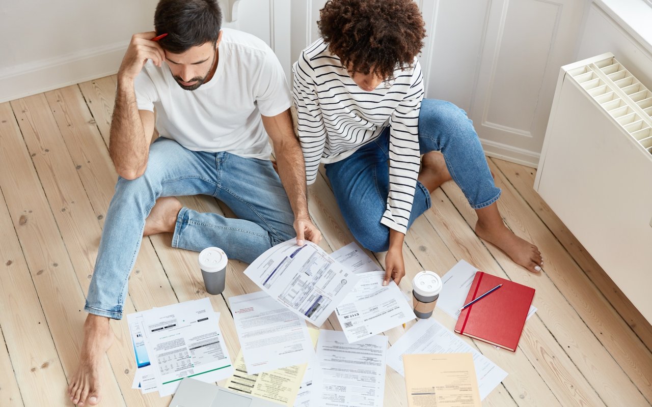 Two adults review scattered paperwork on a wood floor, with coffee cups and a red folder nearby.