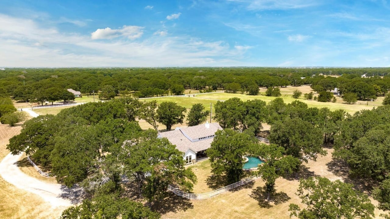 Aerial view of a ranch home and pool surrounded by trees and open land.