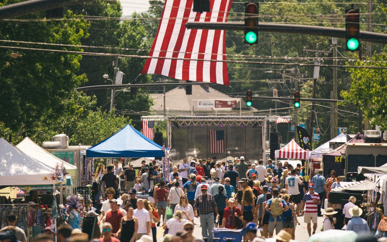Crowds gather at an outdoor festival beneath vendor tents and a large American flag.